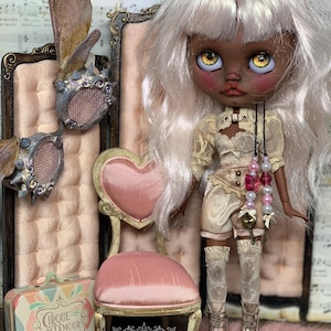 May include: A custom art doll with pale skin, white hair, and yellow eyes, wearing a cream-colored outfit with lace details. The doll is posed with a miniature pink chair and a decorative suitcase. The background includes a pink velvet chair.
