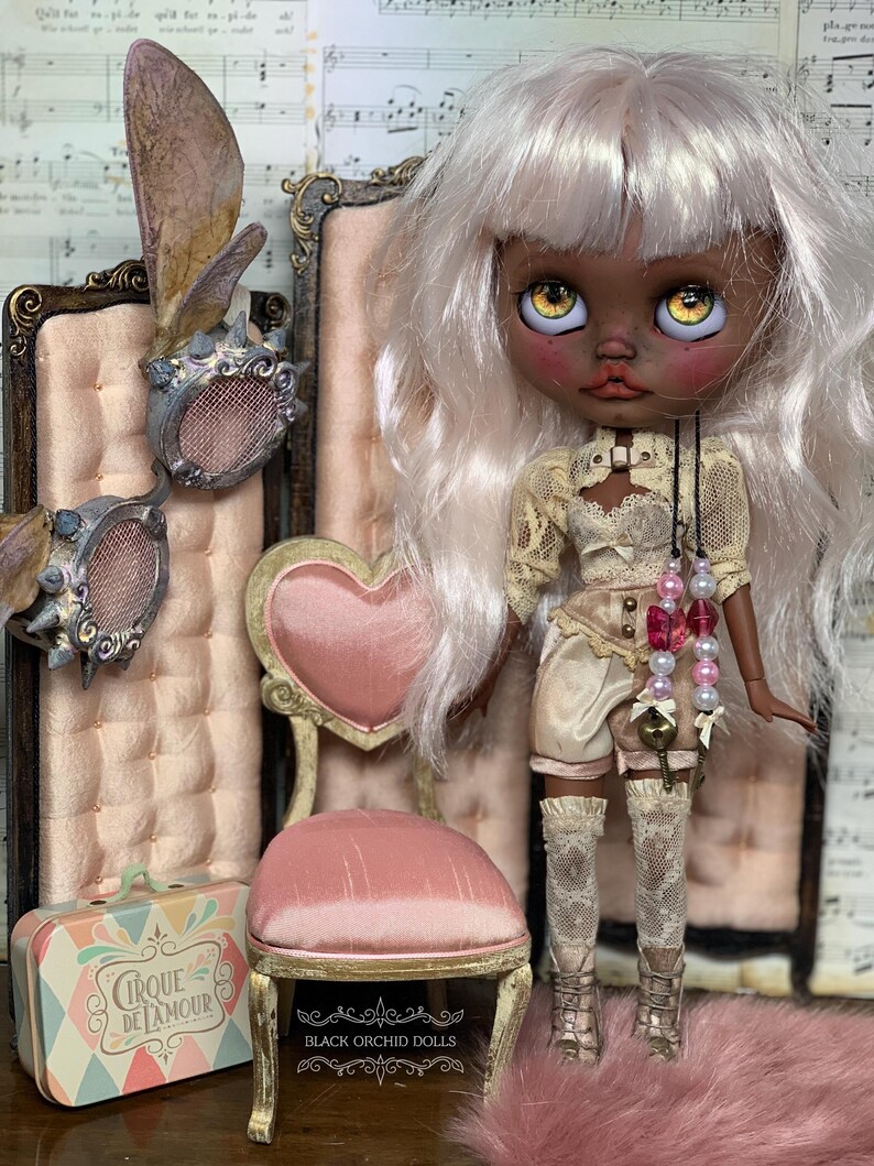 May include: A custom art doll with pale skin, white hair, and yellow eyes, wearing a cream-colored outfit with lace details. The doll is posed with a miniature pink chair and a decorative suitcase. The background includes a pink velvet chair.