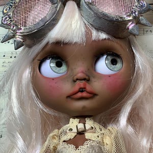 May include: A custom art doll with a detailed face, green eyes, and light pink hair. The doll wears steampunk-style goggles and a cream-colored lace outfit. The text "BLACK ORCHID DOLLS" is visible.