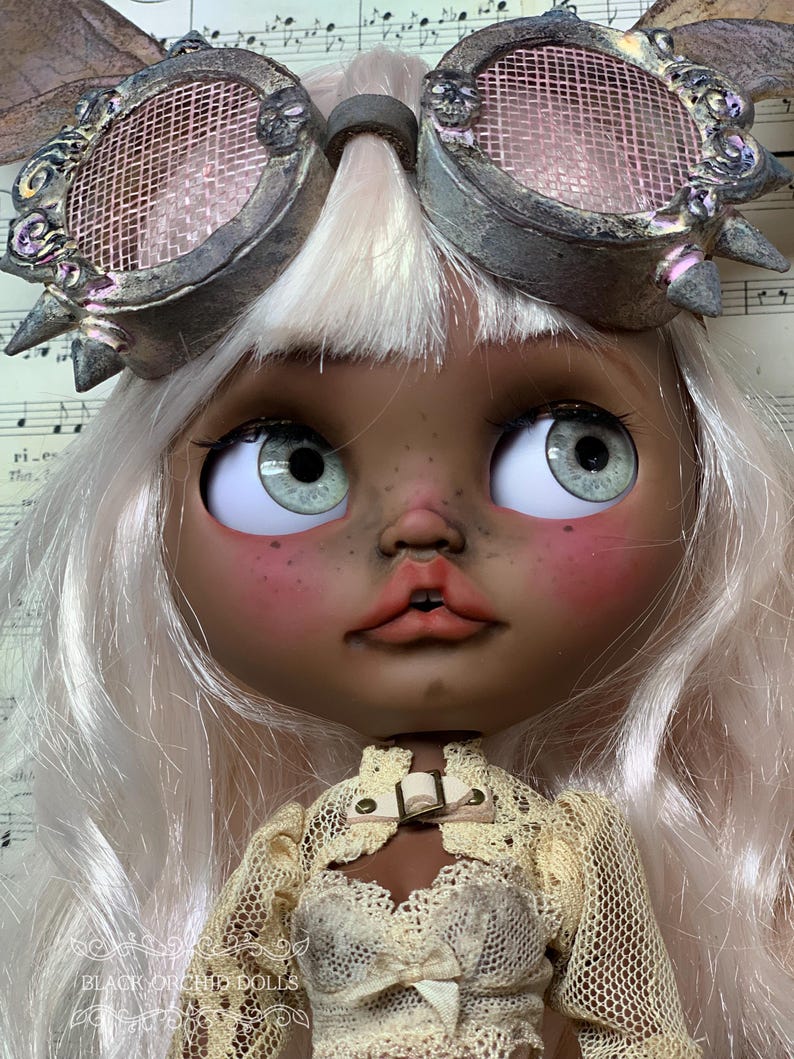 May include: A custom art doll with a detailed face, green eyes, and light pink hair. The doll wears steampunk-style goggles and a cream-colored lace outfit. The text "BLACK ORCHID DOLLS" is visible.