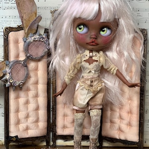 May include: A custom art doll with pale skin, green eyes, and white hair, wearing a cream-colored outfit with lace details and matching boots. The doll stands in front of a pink screen. The text "BLACK ORCHID DOLLS" is visible.