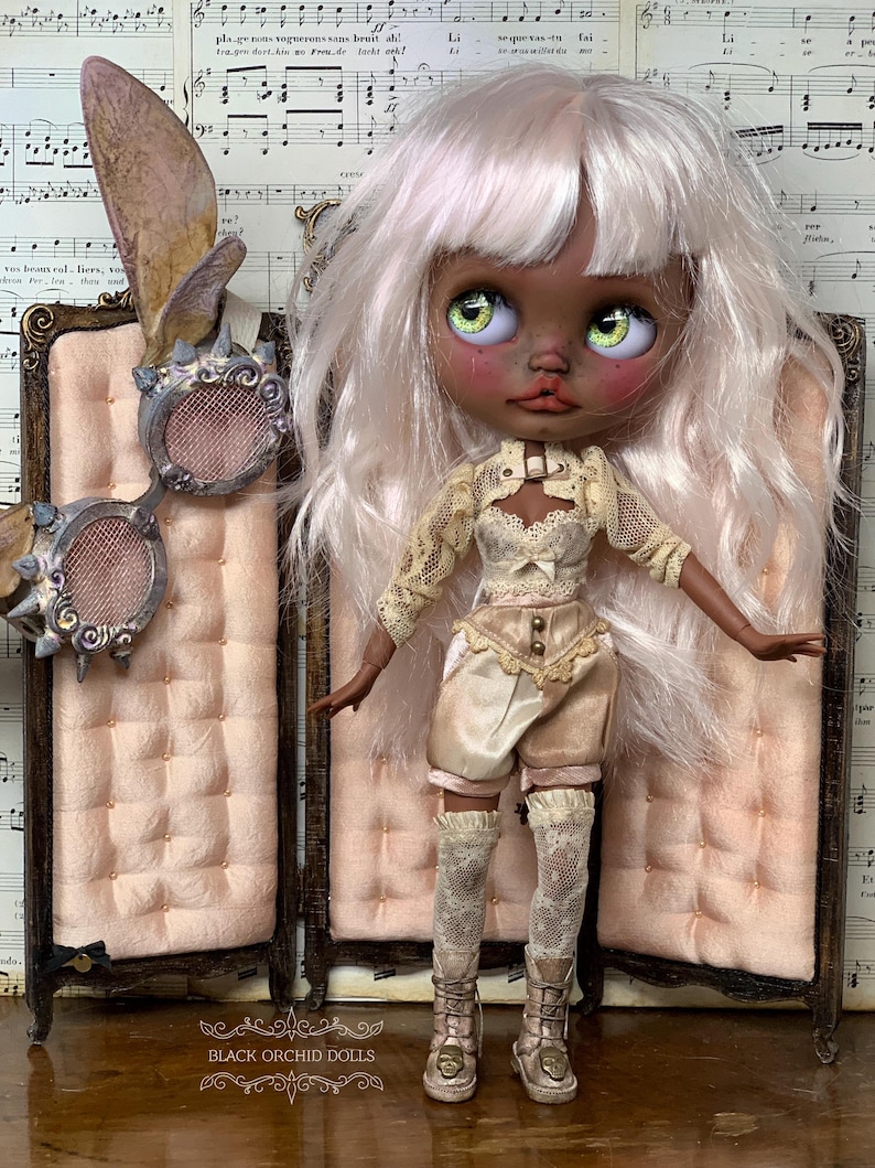 May include: A custom art doll with pale skin, green eyes, and white hair, wearing a cream-colored outfit with lace details and matching boots. The doll stands in front of a pink screen. The text "BLACK ORCHID DOLLS" is visible.