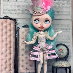 Könnte beinhalten: Puppe mit blauen Haaren und einem rosa Zylinder, gekleidet in einem Outfit im Vintage-Stil. Die Puppe hat einen hellen Teint, Sommersprossen und posiert vor einem Hintergrund aus Notenblättern. Das Outfit umfasst ein Korsett, einen Rock und Strümpfe.