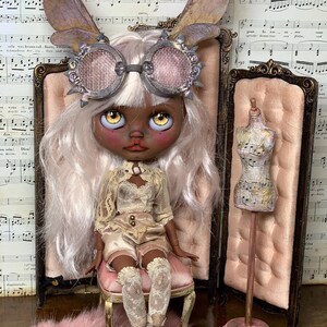 May include: A custom art doll with a dark complexion, wearing a cream-colored outfit, steampunk goggles, and fairy wings. The doll is seated on a pink chair, with a miniature dress form nearby. The background includes sheet music and a pink rug.