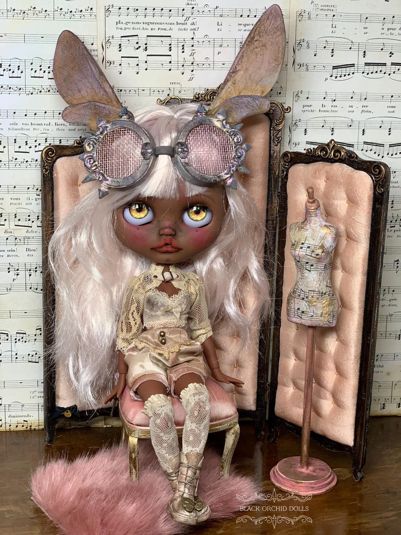 May include: A custom art doll with a dark complexion, wearing a cream-colored outfit, steampunk goggles, and fairy wings. The doll is seated on a pink chair, with a miniature dress form nearby. The background includes sheet music and a pink rug.