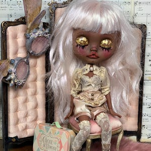 May include: A custom art doll with white hair and gold leaf eyes, wearing a cream-colored outfit and lace stockings. The doll sits on a pink chair next to a small suitcase with the text "Cirque De L'Amour".