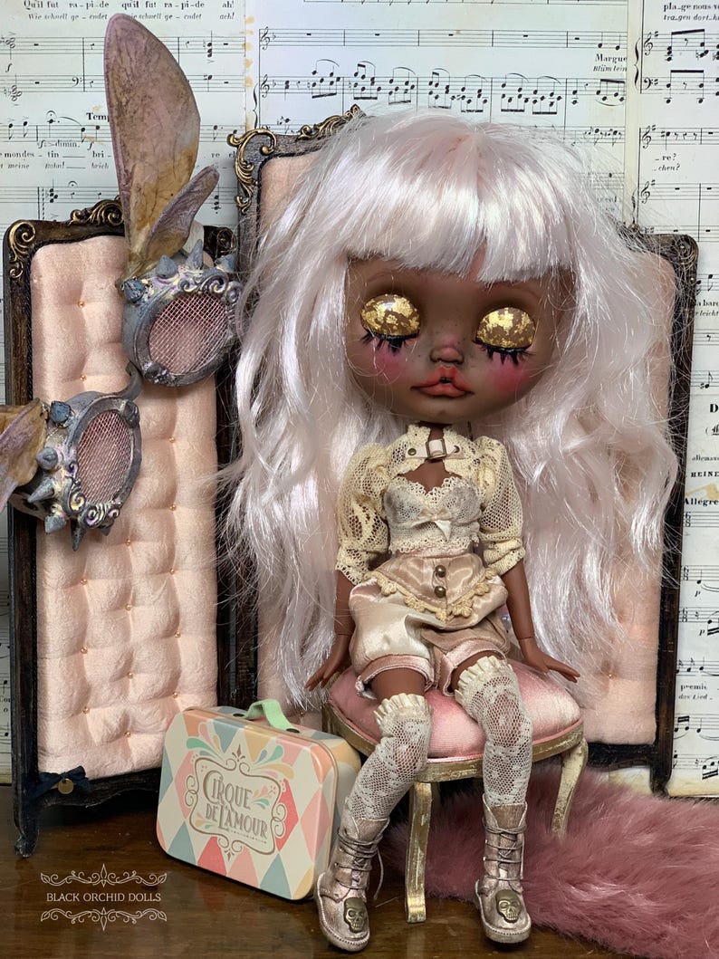 May include: A custom art doll with white hair and gold leaf eyes, wearing a cream-colored outfit and lace stockings. The doll sits on a pink chair next to a small suitcase with the text "Cirque De L'Amour".