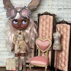 May include: A custom doll with a steampunk aesthetic, wearing goggles, fairy wings, and a detailed outfit. The doll is posed with a pink heart-backed chair, a miniature dress form, and a small suitcase. The background features vintage sheet music.