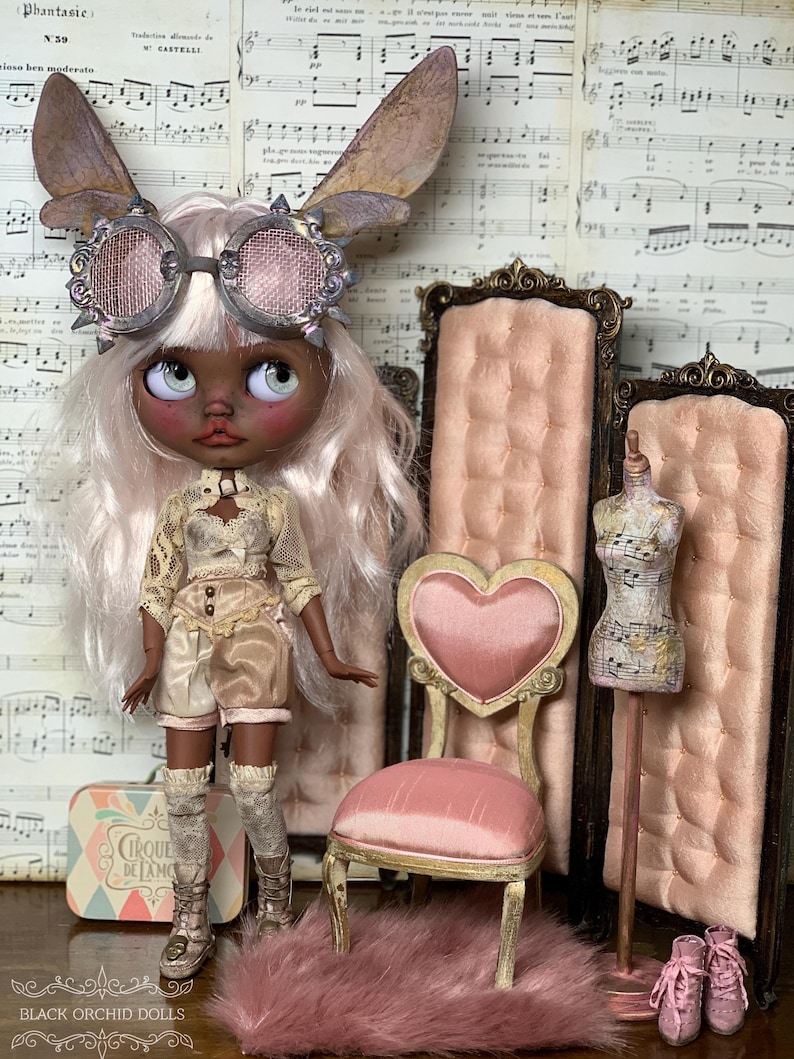 May include: A custom doll with a steampunk aesthetic, wearing goggles, fairy wings, and a detailed outfit. The doll is posed with a pink heart-backed chair, a miniature dress form, and a small suitcase. The background features vintage sheet music.