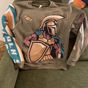 May include: Olive green sweatshirt featuring a warrior design on the front. The warrior is depicted in gold, brown, and blue, holding a shield. The sleeves have a patchwork design with teal, orange, and white accents. The sweatshirt is made of a soft material.