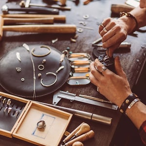 Könnte beinhalten: Eine Person arbeitet an einem Schmuckstück an einem Werktisch. Der Werktisch ist mit Werkzeugen und Zubehör bedeckt, darunter ein Hammer, Zangen, ein Lineal und eine Vielzahl von Ringen und anderen Schmuckstücken. Die Person trägt ein rot-schwarz kariertes Hemd und ein Lederarmband.