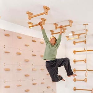 Puede incluir: Una estructura de escalada interior de madera clara que cuenta con una pared con agarres horizontales y un sistema de barras de suspensión montado en el techo. El aparato consta de varias barras cilíndricas y soportes rectangulares de madera.
