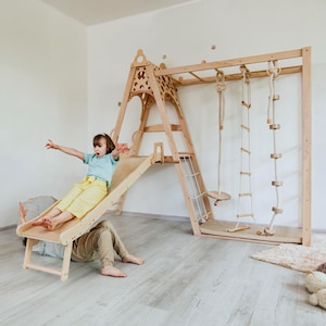 以下が含まれることがあります： 滑り台、クライミングラダー、リング、ロープブランコを備えた木製の屋内プレイセット。明るい色の木材で作られており、子供の遊び用に設計されています。子供が滑り台を滑っており、別の子供が下にいます。