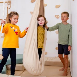 Puede incluir: Tres niños jugando con un columpio de tela beige. El columpio está suspendido del techo. Un niño está dentro del columpio, mientras que los otros dos sostienen los lados. Los niños visten ropa colorida. El fondo incluye presas de escalada y colchonetas.