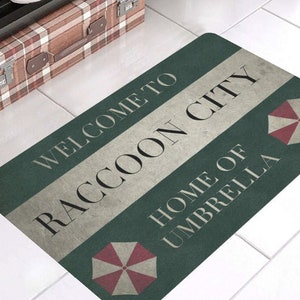 以下が含まれることがあります： 「Welcome to Raccoon City Home of Umbrella」という文字と、赤と白の傘のロゴが書かれた緑と白のウェルカムマットです。