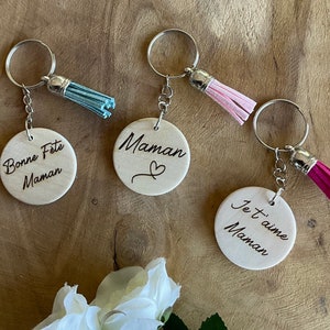 Peut inclure: Trois porte-clés en bois avec des messages gravés différents. Les porte-clés ont des anneaux argentés et des pompons colorés. Les messages sont "Bonne Fete Maman", "Maman" avec un symbole de cœur, et "Je t'aime Maman".