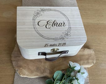 Puede incluir: Una caja de madera de color claro con un asa de cuero marrón oscuro y un cierre de metal. La tapa presenta un diseño de corona floral grabado con láser que rodea el nombre Ebrar y la fecha 25 mars 2024 en una fuente de estilo cursivo.