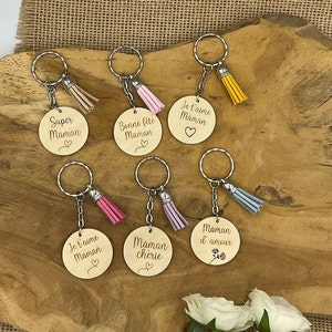 Peut inclure: Sept porte-clés en bois avec différents messages en français, chacun avec un petit pompon. Les porte-clés sont sur une surface en bois. Les messages incluent "Super Maman", "Bonne fête Maman", "Je t'aime Maman", "Je t'aime Maman", "Maman chérie", et "Maman d'amour".