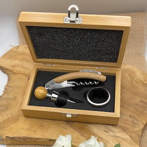May include: A wooden box containing a corkscrew, bottle stopper, and a ring for a wine glass. The box is made of light brown wood and has a black interior.