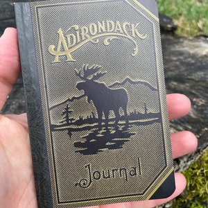 May include: A dark green journal with a gold embossed design featuring the text "Adirondack Journal" and a silhouette of a moose in a forest setting.