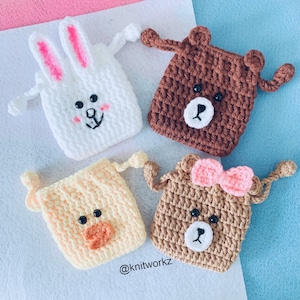May include: Four handmade crochet animal pouches: a white bunny with pink ears, a brown bear, a light yellow duck, and a tan bear with a pink bow. Each pouch has black button eyes and a stitched face. The pouches are on a pink and blue background.