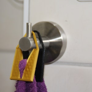 Può includere: Un gancio argentato montato su una parete bianca con un asciugamano viola appeso. L'asciugamano è tenuto in posizione da un gancio giallo stampato in 3D.