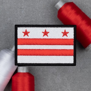 May include: Embroidered patch featuring the flag of Washington, D.C. The design includes three red stars above two red horizontal stripes on a white background, framed by a black border. Red and white spools of thread are in the background.