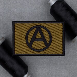 May include: A rectangular patch with a black border and a gold-colored background featuring a black "A" inside a circle. Two spools of black thread are in the background, suggesting a sewing or crafting theme.