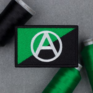 May include: Embroidered patch featuring the Anarchy symbol, a white 'A' inside a circle, set against a green and black background. The patch is rectangular with a black border. Green and black thread spools are in the background.