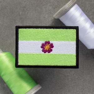 Pode incluir: Etiqueta bordada verde, branca e roxa com um desenho de flor. A etiqueta tem uma borda preta e é feita de tecido.