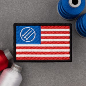 Op de afbeelding: Geborduurde patch met een vlagontwerp. Het blauwe veld heeft een witte cirkel met drie pijlen naar beneden. Rode en witte strepen vormen de vlag, met een zwarte rand. Spoelen met rood, wit en blauw naaigaren staan op de achtergrond.