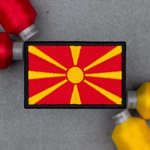 May include: A rectangular embroidered patch featuring the flag of North Macedonia, which consists of a red field with a central yellow sun and eight radiating yellow rays that extend to the edges, all bordered by a thick black stitched frame.