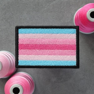 May include: A black framed embroidered patch with a horizontal striped pattern in pink, light pink, and blue. The patch is surrounded by spools of pink thread.