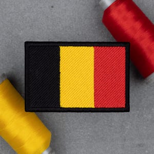 Op de afbeelding: Een rechthoekige patch met de Belgische vlag, met verticale strepen in zwart, geel en rood. De patch heeft een zwarte rand en is geplaatst tegen een grijze achtergrond. Twee garenklosjes, één rood en één geel, zijn zichtbaar.