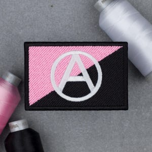 May include: Embroidered patch featuring a pink and black design with a white encircled letter "A". The patch is rectangular with a black border. Spools of thread in pink, white, and black are in the background.
