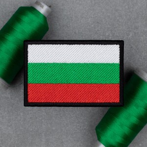 May include: Embroidered patch featuring the flag of Bulgaria. The rectangular patch has three horizontal stripes in white, green, and red, outlined with a black border. Two spools of green thread are in the background.