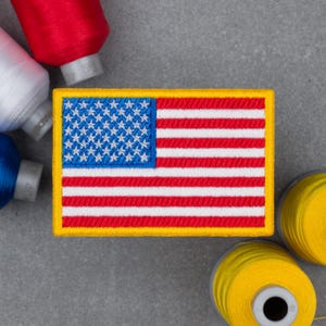 May include: Embroidered patch featuring the American flag with red and white stripes and a blue field of white stars. The patch has a yellow border. Spools of red, white, blue, and yellow thread are arranged around the patch on a gray surface.