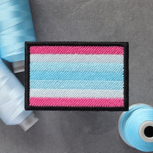 May include: A blue, white, and pink transgender pride flag patch with black stitching around the edges.