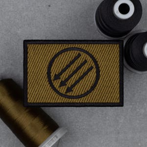Puede incluir: Parche rectangular con un diseño dorado y negro. El parche presenta un círculo negro con tres flechas que apuntan hacia abajo en el interior. El parche está rodeado por un borde negro y tiene una apariencia texturizada. Los carretes de hilo de coser están en el fondo.