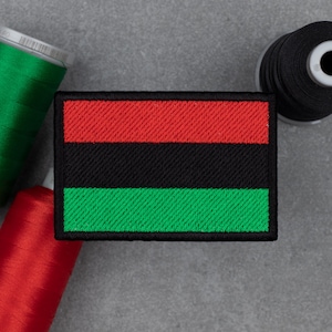 May include: A rectangular patch featuring a red, black, and green striped design, with a black border. Spools of red, green, and black thread are arranged around the patch on a grey surface.