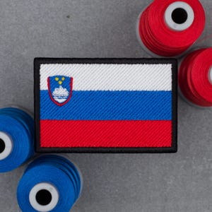 Puede incluir: Un parche rectangular bordado que muestra la bandera de Eslovenia con franjas horizontales blanca, azul y roja y un escudo en la esquina superior izquierda, rodeado por varios carretes de hilo de coser rojo y azul.