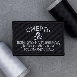 May include: A black rectangular patch with white embroidered text and a skull and crossbones symbol. The text reads "СМЕРТЬ" above and a longer phrase below. Two spools of thread, one black and one white, are in the background.