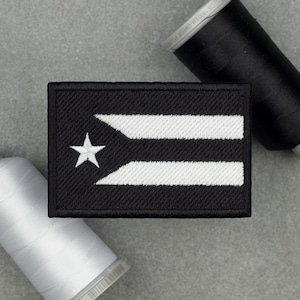 Puede incluir: Un parche rectangular bordado en blanco y negro con la bandera de Puerto Rico. El parche tiene un fondo negro con una estrella blanca y dos franjas blancas. El parche está rodeado por un borde negro.