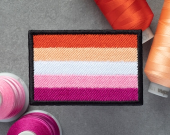 May include: A lesbian pride flag patch with six horizontal stripes in the colors orange, light peach, white, light pink, pink, and dark purple. The patch is surrounded by a black border.
