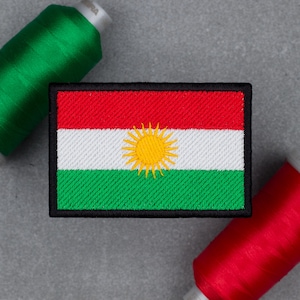 Può includere: Toppa ricamata raffigurante la bandiera del Kurdistan, con strisce orizzontali rosse, bianche e verdi e un emblema solare giallo centrale. La toppa è rettangolare con un bordo nero. Sullo sfondo ci sono rocchetti di filo verde e rosso.