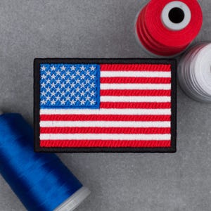 Op de afbeelding: Geborduurde patch met de Amerikaanse vlag, met rode en witte strepen en een blauw veld met witte sterren, omlijst door een zwarte rand. Spoelen met rood, wit en blauw garen zijn op de achtergrond.