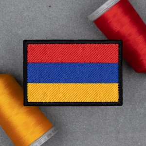 May include: Embroidered patch featuring the flag of Armenia. The rectangular patch has a black border and displays the flag's horizontal stripes in red, blue, and yellow. Red and yellow spools of thread are visible in the background.