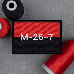 Könnte beinhalten: Rechteckiger, gestickter Aufnäher in Rot und Schwarz. Der obere Teil ist rot, der untere Teil ist schwarz, mit dem weißen Text "M-26-7". Spulen mit schwarzem und rotem Faden sind im Hintergrund zu sehen.