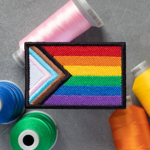 May include: A rainbow pride flag with a brown, blue, light blue, and pink triangle in the corner. The flag is embroidered and surrounded by spools of thread in pink, blue, yellow, and orange.
