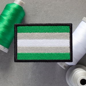 Puede incluir: Un parche bordado que representa la bandera del orgullo genderqueer. El parche es verde, blanco y gris con un borde negro.